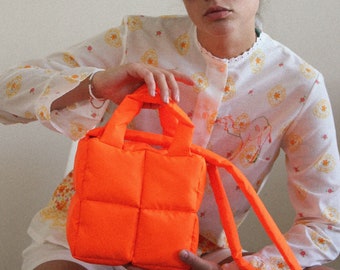 Könnte beinhalten: Leuchtend orangefarbene, gesteppte Tragetasche mit kurzen Griffen und einem langen Riemen. Die Tasche ist quadratisch und gepolstert. Die Person im Bild trägt ein weißes Oberteil mit Blumenmuster und hellblaue Sonnenbrillen.