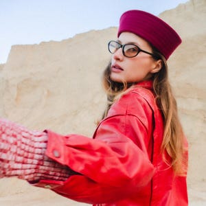 May include: A bright red jacket with a matching maroon hat and black-framed glasses. The jacket has a classic bomber style. The sleeves are cuffed, and the jacket is paired with a white, open-weave garment.