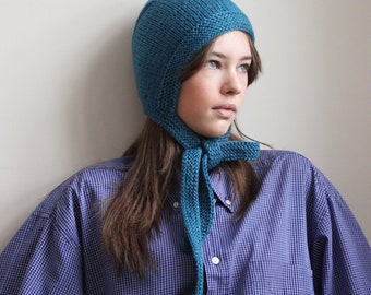 May include: A teal blue knitted hat with a long scarf attached. The hat has a pointed crown and a flap that covers the ears. The scarf is tied in a loose knot at the neck. The person wearing the hat is wearing a blue and white plaid shirt.