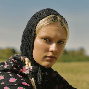 Può includere: Una donna con i capelli biondi indossa un berretto nero a maglia e un vestito nero a fiori. Il berretto ha un design a uncinetto e le copre le orecchie. Sta guardando la telecamera.