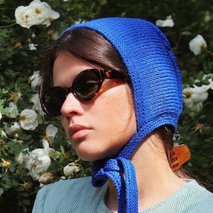Könnte beinhalten: Eine Frau trägt eine blaue Strickmütze mit Schleife, Sonnenbrille und ein hellgrünes, gestreiftes Hemd. Sie steht vor einem Busch mit weißen Blüten.