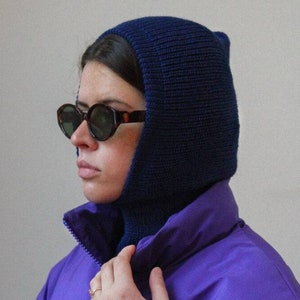May include: A person wearing a purple puffer jacket with a blue knitted balaclava and tortoise shell sunglasses.