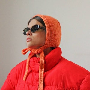 Könnte beinhalten: Eine leuchtend orangefarbene Strickmütze mit langem Band. Die Mütze wird von einer Person getragen, die eine rote Daunenjacke und eine schwarze Sonnenbrille trägt.