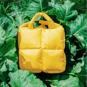 May include: A bright yellow quilted tote bag with a single handle. The bag is sitting on a bed of green leaves.