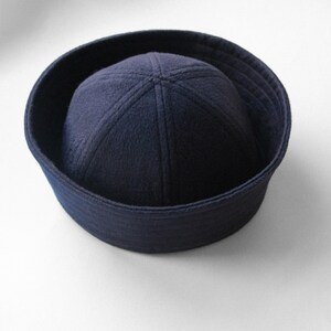 May include: A navy blue sailor hat with a rounded crown and a flat, circular brim. The hat is made of a soft, felt-like material and is a classic nautical accessory. The hat's brim measures approximately 10 inches in diameter.