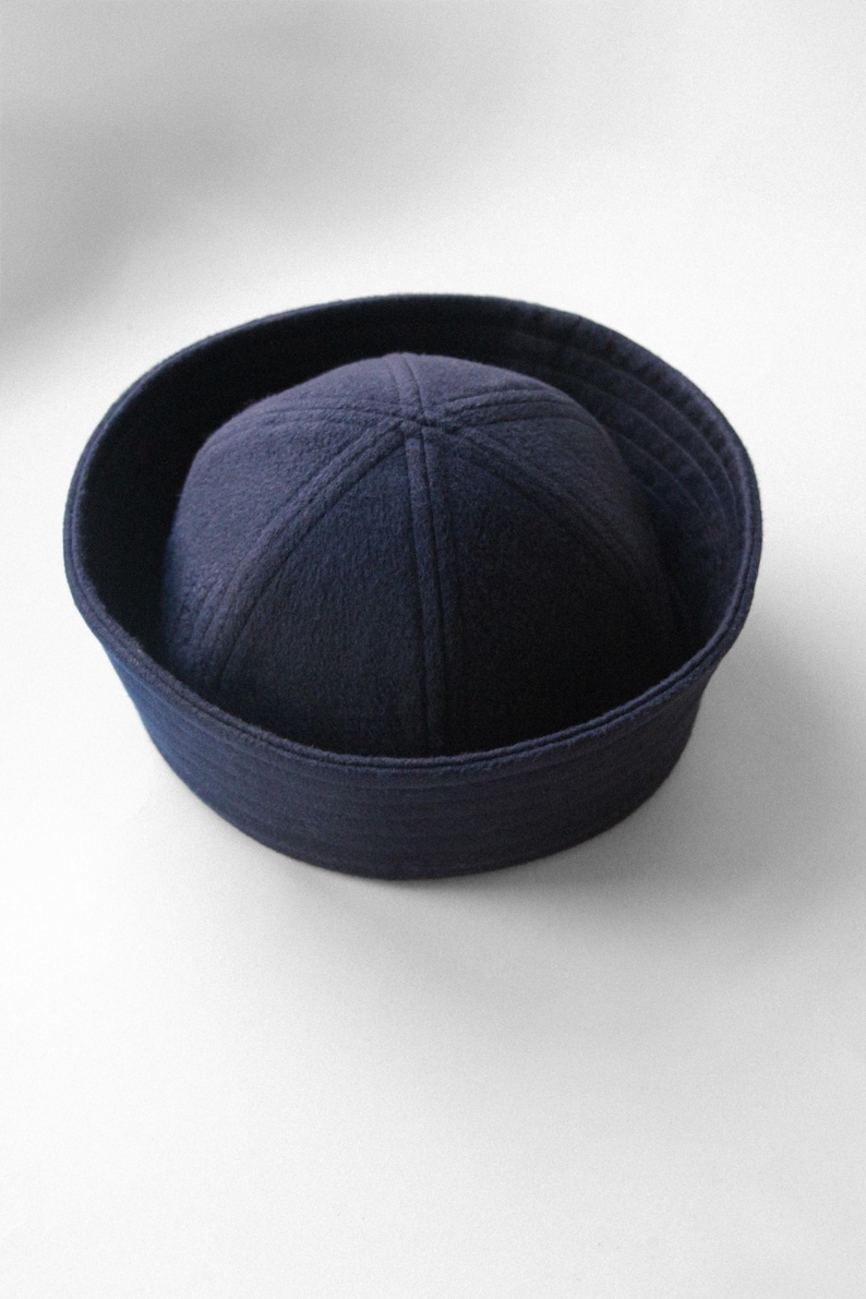 May include: A navy blue sailor hat with a rounded crown and a flat, circular brim. The hat is made of a soft, felt-like material and is a classic nautical accessory. The hat's brim measures approximately 10 inches in diameter.