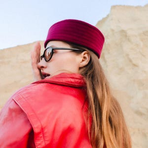 May include: A person wearing a magenta-colored hat and black-framed sunglasses. The person is also wearing a red jacket. The background is a light tan color.
