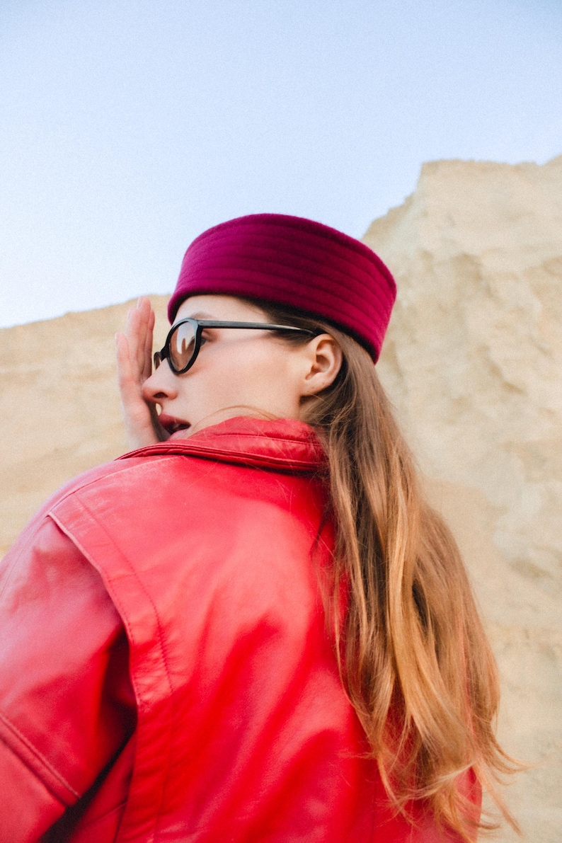 May include: A person wearing a magenta-colored hat and black-framed sunglasses. The person is also wearing a red jacket. The background is a light tan color.