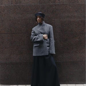 May include: A grey pinstriped blazer with a high collar and long sleeves, paired with a long black skirt, a black hat, and black shoes. The blazer features a structured fit with two front pockets and a vertical white pinstripe pattern.