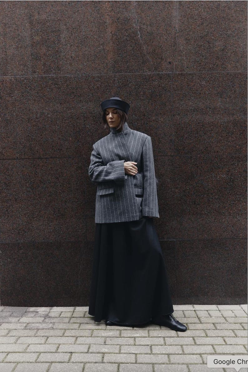 May include: A grey pinstriped blazer with a high collar and long sleeves, paired with a long black skirt, a black hat, and black shoes. The blazer features a structured fit with two front pockets and a vertical white pinstripe pattern.