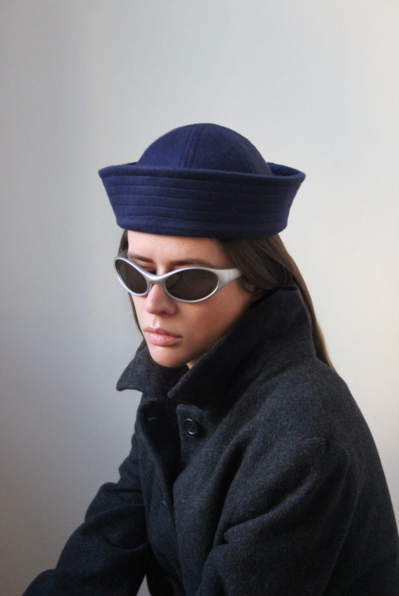 May include: A navy blue sailor hat with a structured brim. The hat is worn with silver-framed sunglasses and a dark gray coat. The image is set against a plain, light gray background.