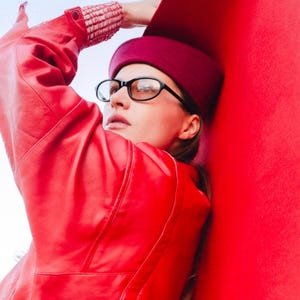 May include: A person wearing a red leather jacket, a burgundy hat, and black-framed glasses. The jacket has visible stitching and a pocket. The background is a solid red color, with a light blue sky.