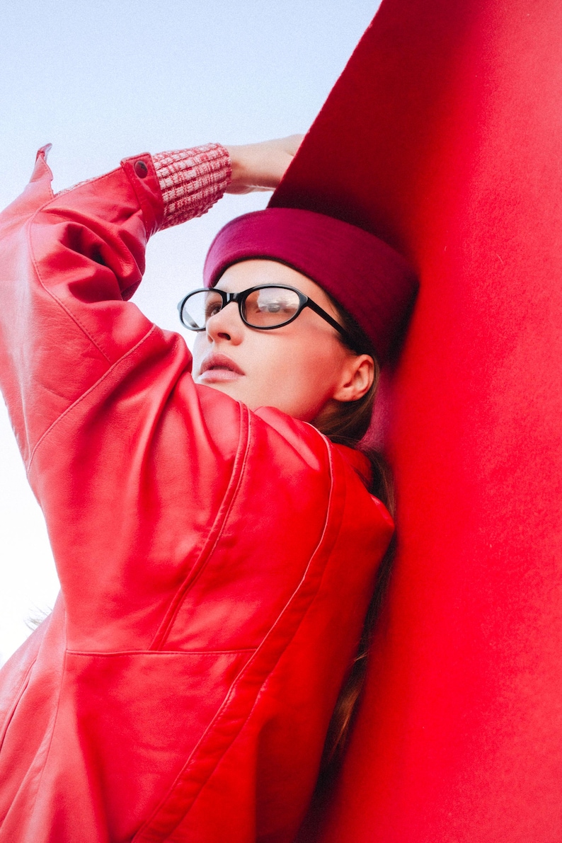May include: A person wearing a red leather jacket, a burgundy hat, and black-framed glasses. The jacket has visible stitching and a pocket. The background is a solid red color, with a light blue sky.