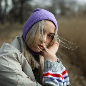 Puede incluir: Una mujer con un gorro morado y una chaqueta beige con una manga a rayas. Su cabello rubio se mueve con el viento.