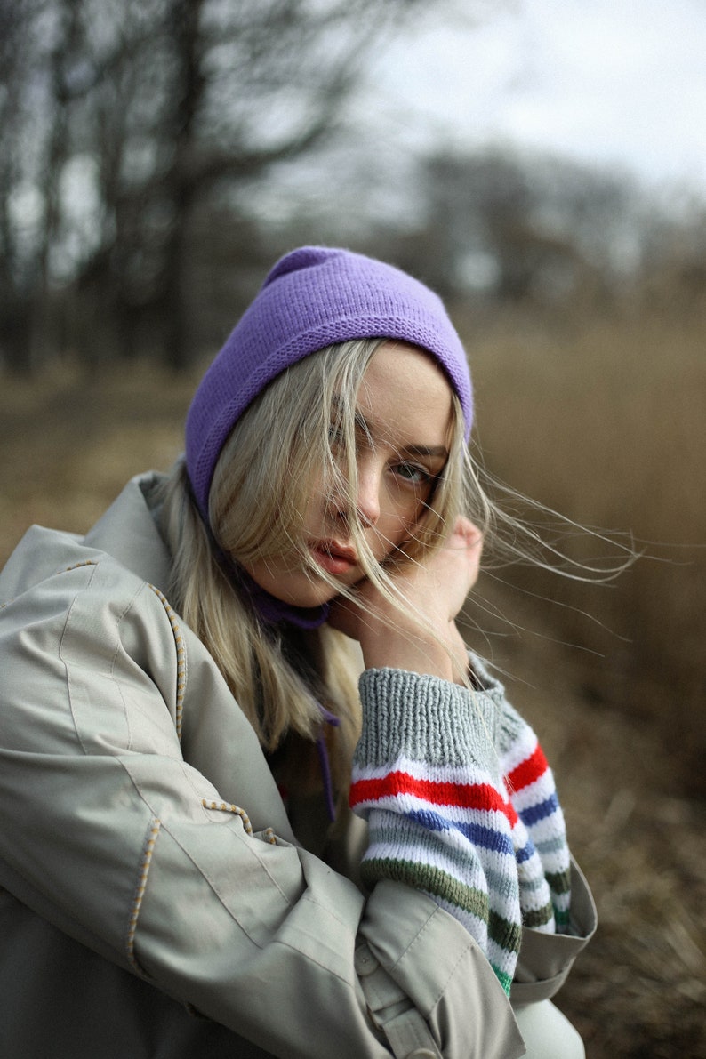 Puede incluir: Una mujer con un gorro morado y una chaqueta beige con una manga a rayas. Su cabello rubio se mueve con el viento.