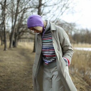 Puede incluir: Una mujer lleva un abrigo beige claro con un gorro morado y un su&eacute;ter a rayas. El abrigo tiene un dise&ntilde;o largo y fluido y se lleva sobre un su&eacute;ter a rayas. La mujer est&aacute; de pie en una zona arbolada con un camino en el fondo.