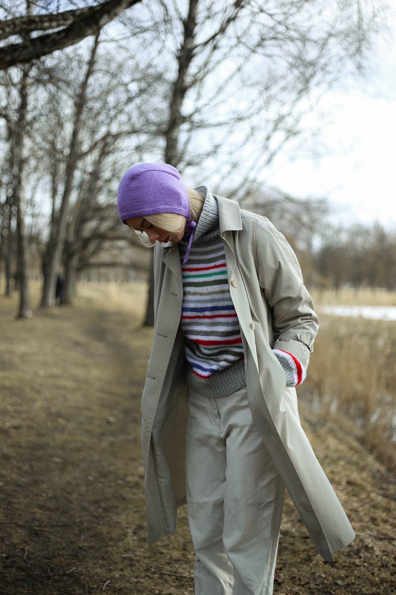 Puede incluir: Una mujer lleva un abrigo beige claro con un gorro morado y un su&eacute;ter a rayas. El abrigo tiene un dise&ntilde;o largo y fluido y se lleva sobre un su&eacute;ter a rayas. La mujer est&aacute; de pie en una zona arbolada con un camino en el fondo.