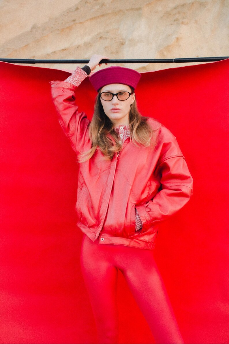 May include: A person wearing a red jacket, leggings, and a beret. The jacket has a zipper closure and pockets. The beret and leggings are a matching shade of red. The person is also wearing glasses.