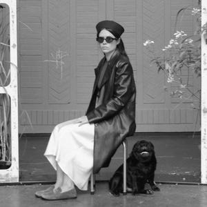 May include: A black leather coat worn over a long white skirt, paired with a black beret and dark sunglasses. A small black pug sits on the floor next to the stool. The ensemble is completed with grey suede loafers, creating a monochromatic look.