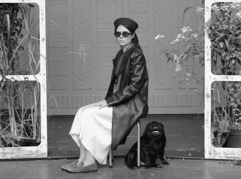 May include: A black leather coat worn over a long white skirt, paired with a black beret and dark sunglasses. A small black pug sits on the floor next to the stool. The ensemble is completed with grey suede loafers, creating a monochromatic look.