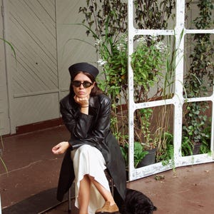 May include: A black beret, black sunglasses, a black leather trench coat, a white midi skirt, and tan suede loafers. The outfit is styled with a long-sleeved black leather coat worn over a flowing white skirt, paired with classic tan suede loafers.