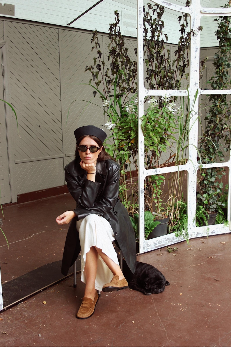 May include: A black beret, black sunglasses, a black leather trench coat, a white midi skirt, and tan suede loafers. The outfit is styled with a long-sleeved black leather coat worn over a flowing white skirt, paired with classic tan suede loafers.