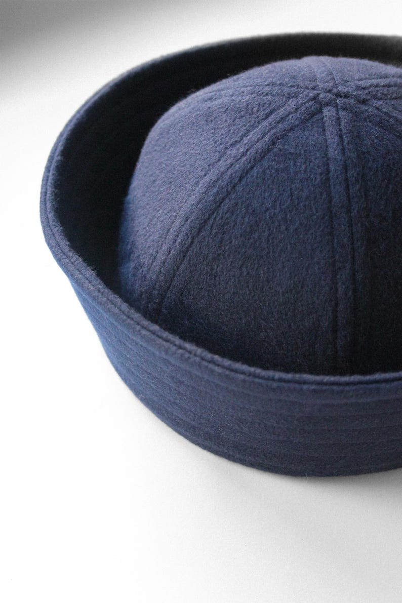 May include: A navy blue sailor hat with a rounded crown and a flat brim. The hat is made of a soft, felt-like material and features stitched details on the crown. The hat is a classic style, often associated with nautical themes.