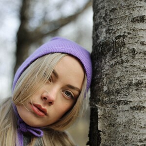 Puede incluir: Una mujer con cabello rubio lleva un gorro y una bufanda de punto morado. Se apoya en un abedul.