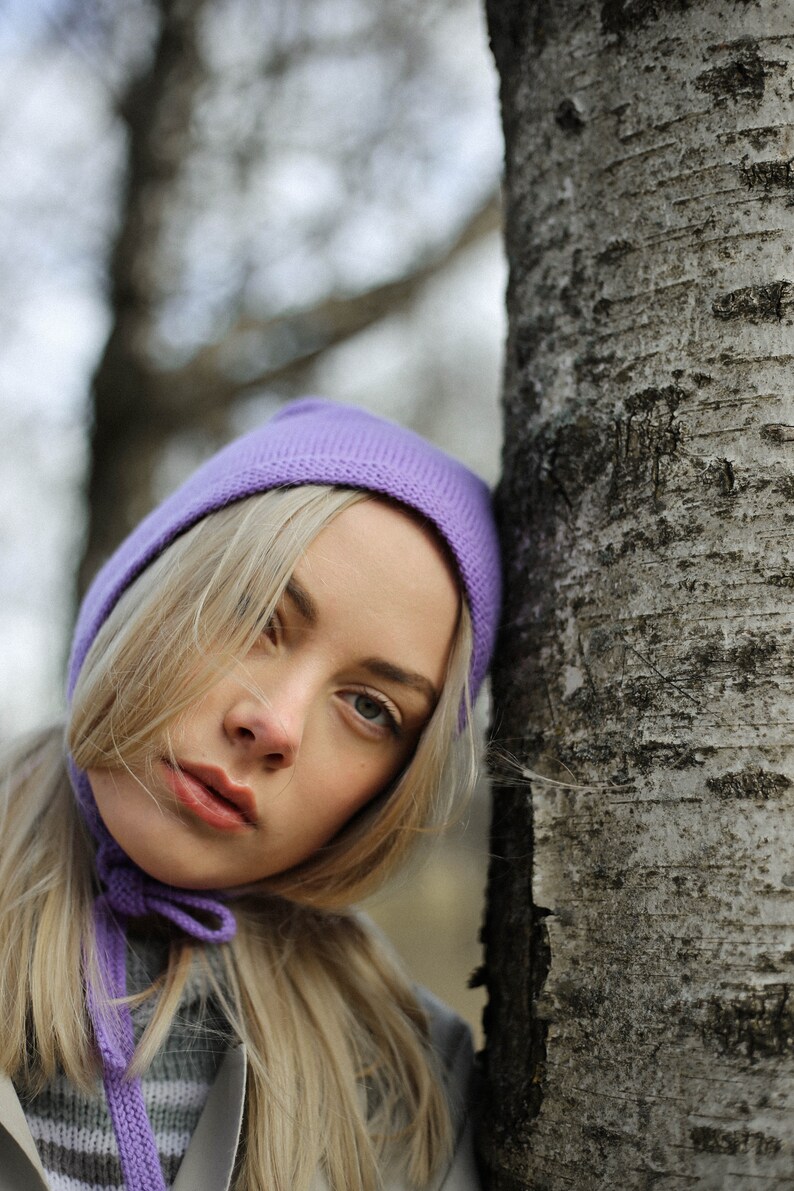 Puede incluir: Una mujer con cabello rubio lleva un gorro y una bufanda de punto morado. Se apoya en un abedul.