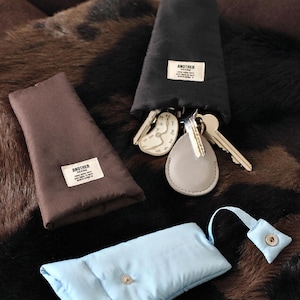 May include: A collection of key pouches and accessories in various colors. There is a black pouch with keys and a gray key fob, a brown pouch, and a light blue pouch with a snap closure. Each pouch has a white label that says "ANOTHER".