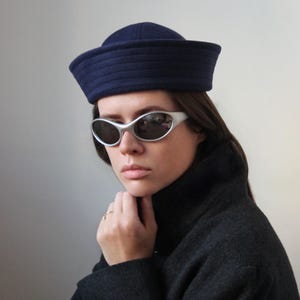May include: A woman wearing a navy blue sailor hat and silver-framed sunglasses. She is also wearing a dark gray coat. The hat has a structured, rounded crown and a flat brim. The sunglasses have a curved, oval shape.