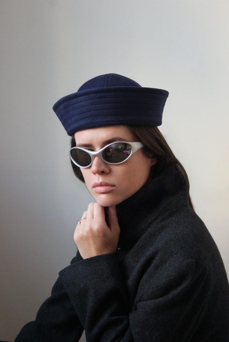May include: A woman wearing a navy blue sailor hat and silver-framed sunglasses. She is also wearing a dark gray coat. The hat has a structured, rounded crown and a flat brim. The sunglasses have a curved, oval shape.