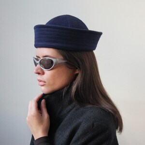 May include: A navy blue sailor hat with a flat top and a structured brim. The hat is worn with silver-framed sunglasses and a dark gray coat. The overall aesthetic is vintage-inspired and chic.