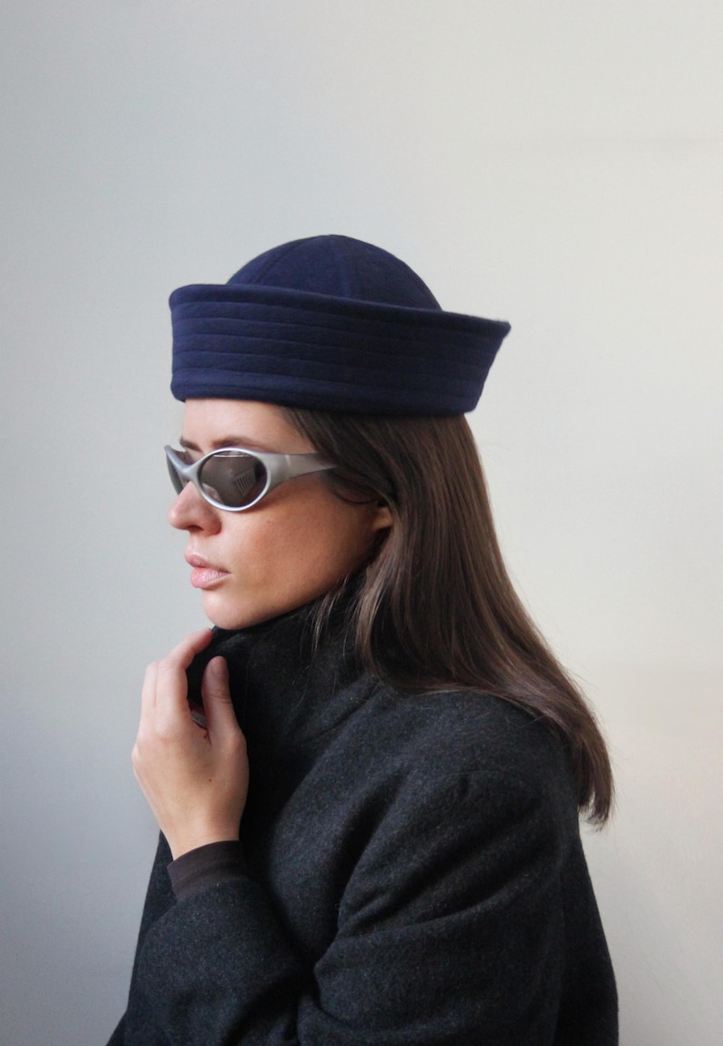 May include: A navy blue sailor hat with a flat top and a structured brim. The hat is worn with silver-framed sunglasses and a dark gray coat. The overall aesthetic is vintage-inspired and chic.