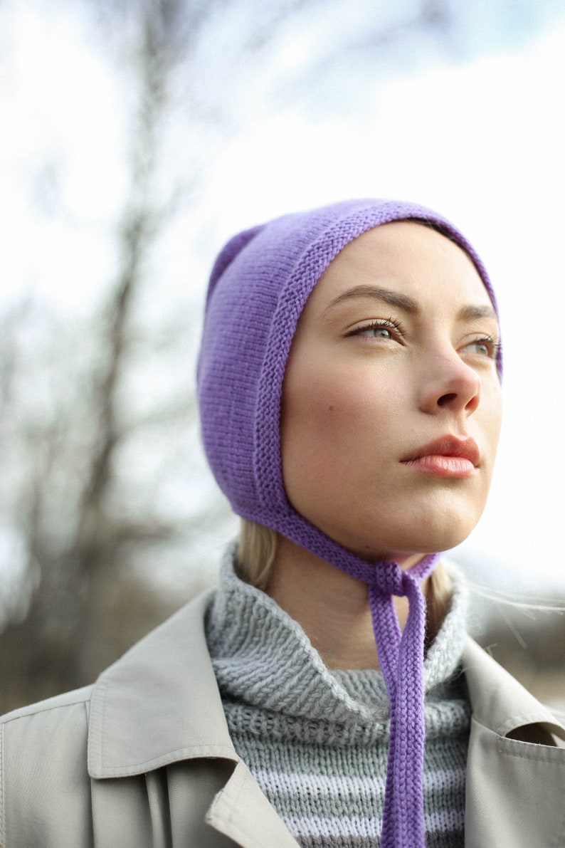 Puede incluir: Una mujer con un gorro de punto morado con una larga corbata. El gorro est&aacute; tirado sobre su frente y la corbata est&aacute; atada alrededor de su cuello. Lleva un su&eacute;ter a rayas grises y blancas y un abrigo beige.