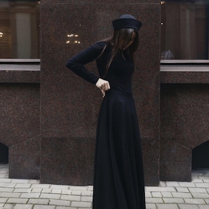 May include: A long-sleeved black top and a matching floor-length black skirt, paired with a black pillbox-style hat. The ensemble features a solid black color throughout, with the skirt having a full, flowing silhouette and the top being fitted.
