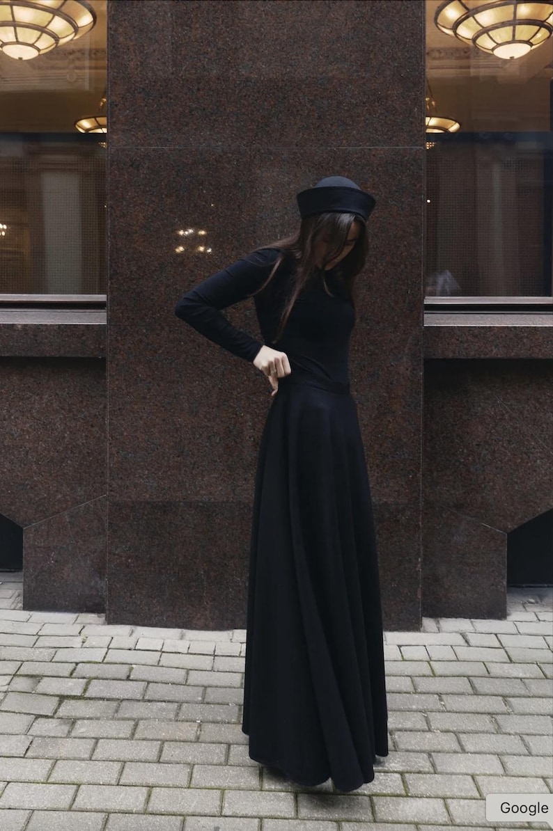 May include: A long-sleeved black top and a matching floor-length black skirt, paired with a black pillbox-style hat. The ensemble features a solid black color throughout, with the skirt having a full, flowing silhouette and the top being fitted.