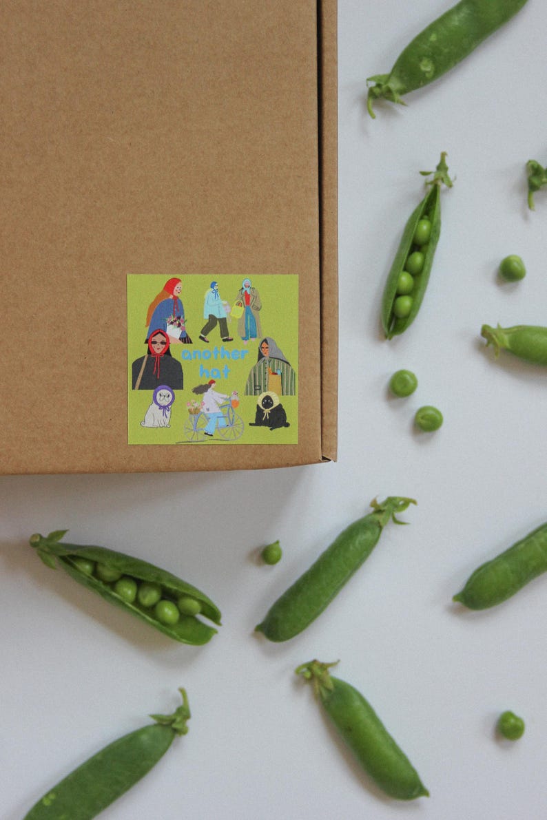 May include: A brown cardboard box with a sticker featuring illustrations of people and the text "another hat." Scattered around are fresh green pea pods and loose peas on a white surface.