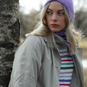 Puede incluir: Una mujer con un gorro de punto de color lila claro, una bufanda larga de color blanco y un su&eacute;ter a rayas. Lleva una chaqueta beige y tiene el pelo largo y rubio.