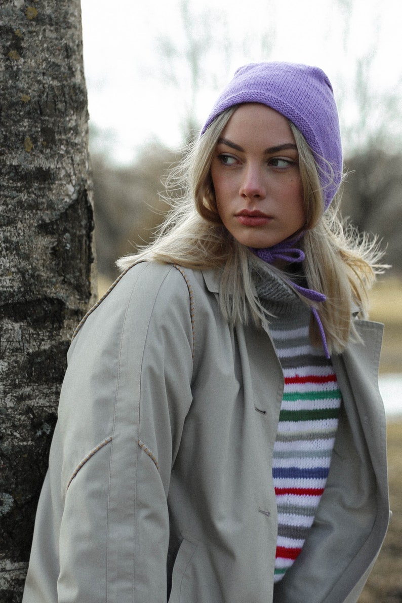 Puede incluir: Una mujer con un gorro de punto de color lila claro, una bufanda larga de color blanco y un su&eacute;ter a rayas. Lleva una chaqueta beige y tiene el pelo largo y rubio.