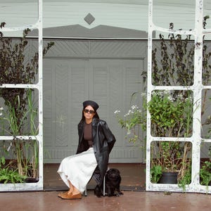 May include: A black beret, dark sunglasses, a brown top, a black leather coat, and a white skirt. The outfit is paired with tan suede loafers. A small black dog sits on the ground nearby, positioned in front of a light green wooden structure.
