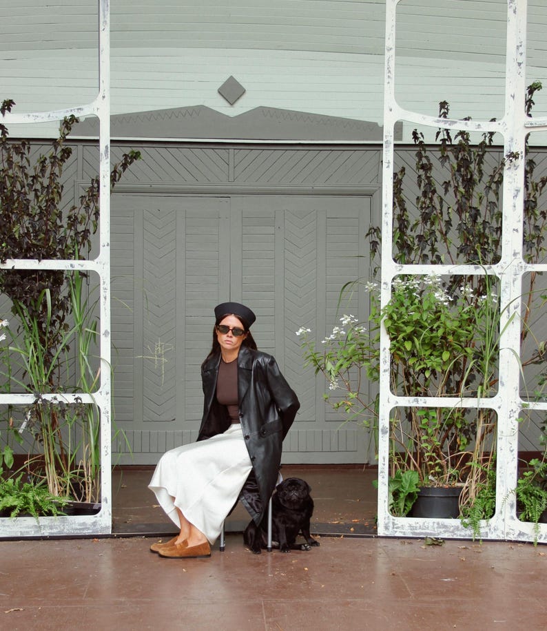 May include: A black beret, dark sunglasses, a brown top, a black leather coat, and a white skirt. The outfit is paired with tan suede loafers. A small black dog sits on the ground nearby, positioned in front of a light green wooden structure.