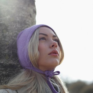 Puede incluir: Una mujer con cabello rubio lleva un gorro de punto morado con un lazo. El gorro est&aacute; atado debajo de la barbilla. Lleva un su&eacute;ter a rayas grises y blancas y una chaqueta beige.