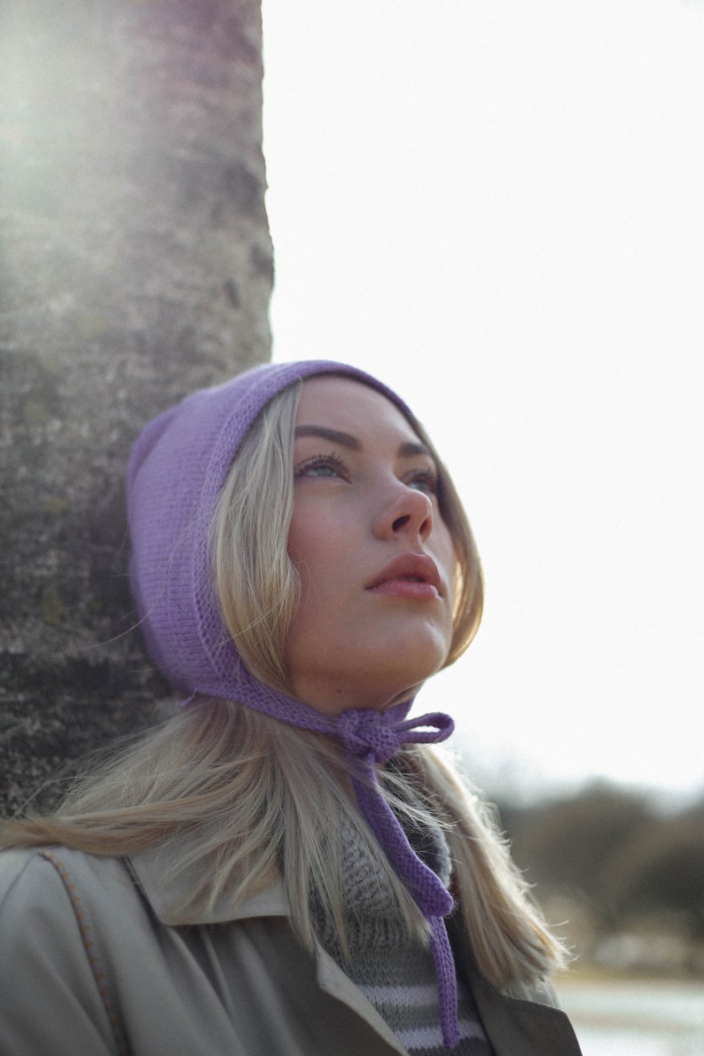 Puede incluir: Una mujer con cabello rubio lleva un gorro de punto morado con un lazo. El gorro est&aacute; atado debajo de la barbilla. Lleva un su&eacute;ter a rayas grises y blancas y una chaqueta beige.