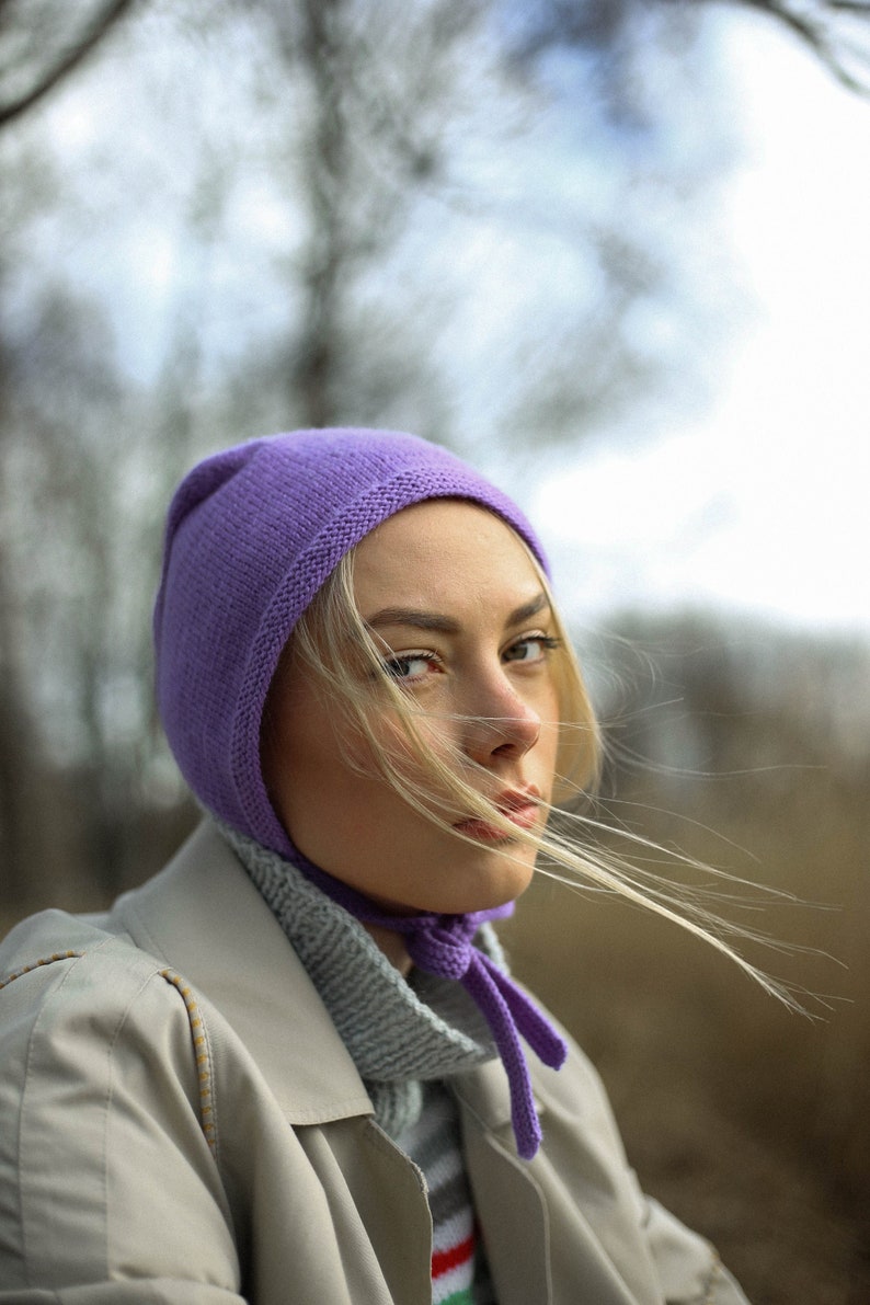 Puede incluir: Una mujer con un gorro de punto morado y una larga cola de caballo rubia. Lleva un abrigo beige y un jersey de cuello alto gris claro.