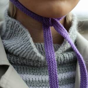 Puede incluir: Primer plano de una persona que lleva una gorra de punto morada atada alrededor del cuello. La gorra est&aacute; atada en un lazo y la persona lleva un su&eacute;ter a rayas gris y blanco.