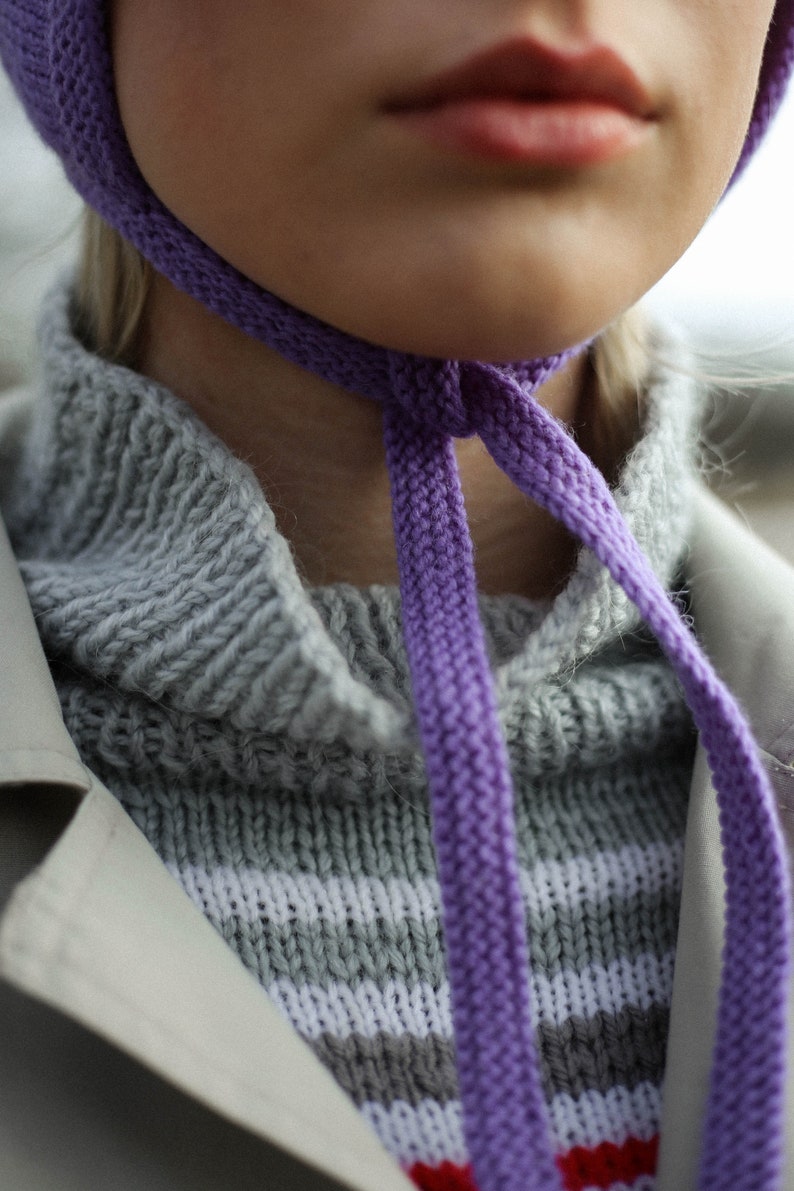 Puede incluir: Primer plano de una persona que lleva una gorra de punto morada atada alrededor del cuello. La gorra est&aacute; atada en un lazo y la persona lleva un su&eacute;ter a rayas gris y blanco.