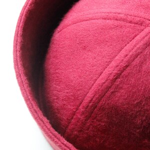 May include: Close-up of a burgundy-colored hat with a rounded crown and a slightly upturned brim. The hat has a textured, felt-like appearance and is likely made of wool or a similar material. The hat's design features stitched seams.