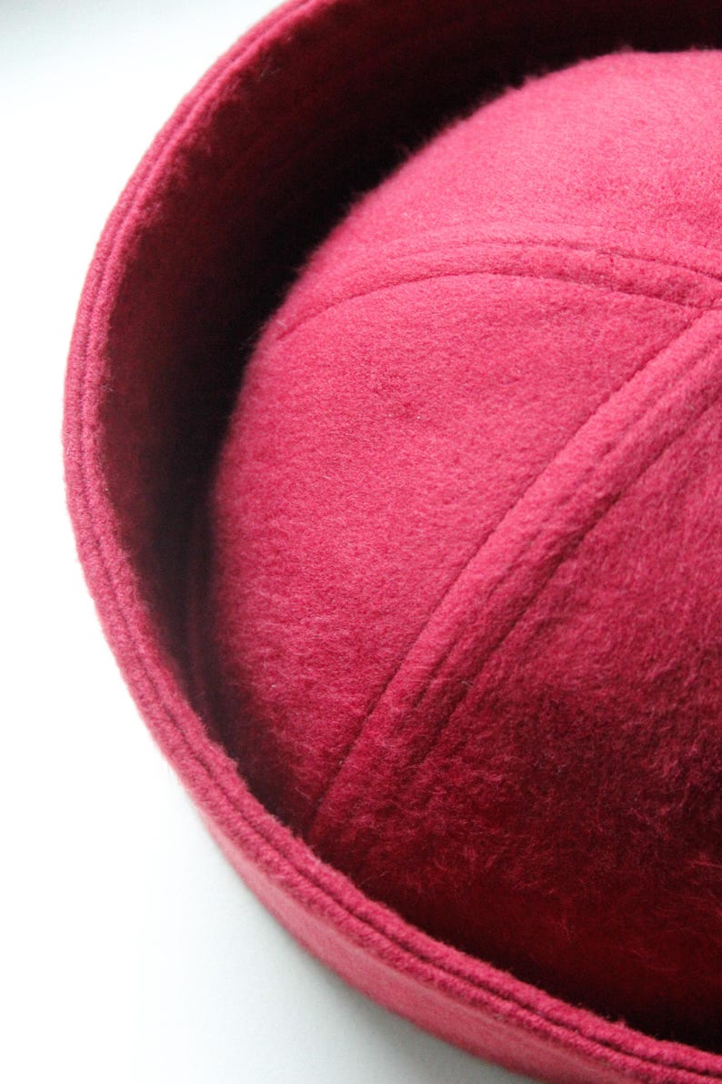 May include: Close-up of a burgundy-colored hat with a rounded crown and a slightly upturned brim. The hat has a textured, felt-like appearance and is likely made of wool or a similar material. The hat's design features stitched seams.