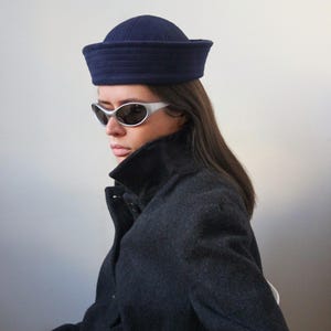 May include: A navy blue sailor hat with a structured brim. The hat is worn with a dark gray coat and silver-framed sunglasses. The hat's design is a classic style, suitable for various outfits.